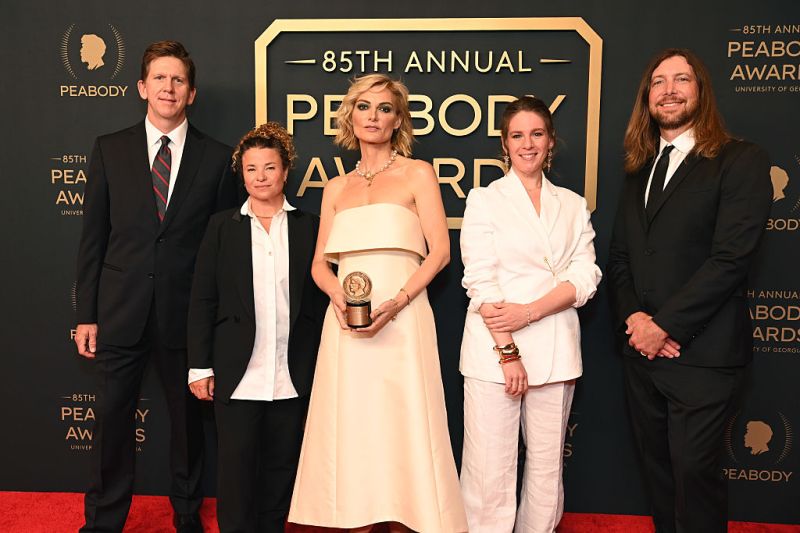 Woman in ivory colored satin gown holding award statuette with two people on either side wearing black and white formal attire standing on a red carpet in front of a sign that says 85th Annual Peabody Awards