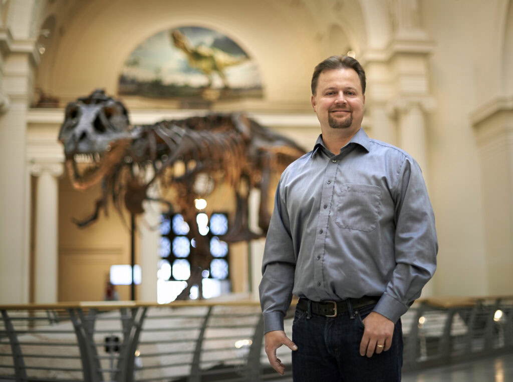 A man in a blue button down shirt stands in front of a T. rex skeleton displayed in the atrium of the Field Museum of Natural History
