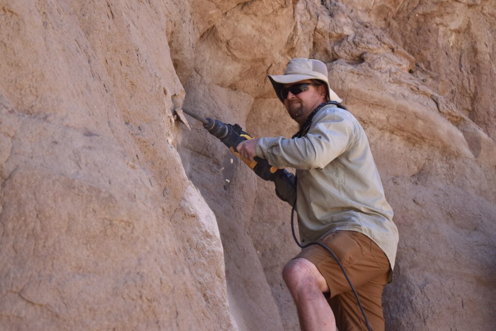 A man wearing a hat and sunglasses is standing on a ledge on a rock face,  using a jackhammer on the rock surface