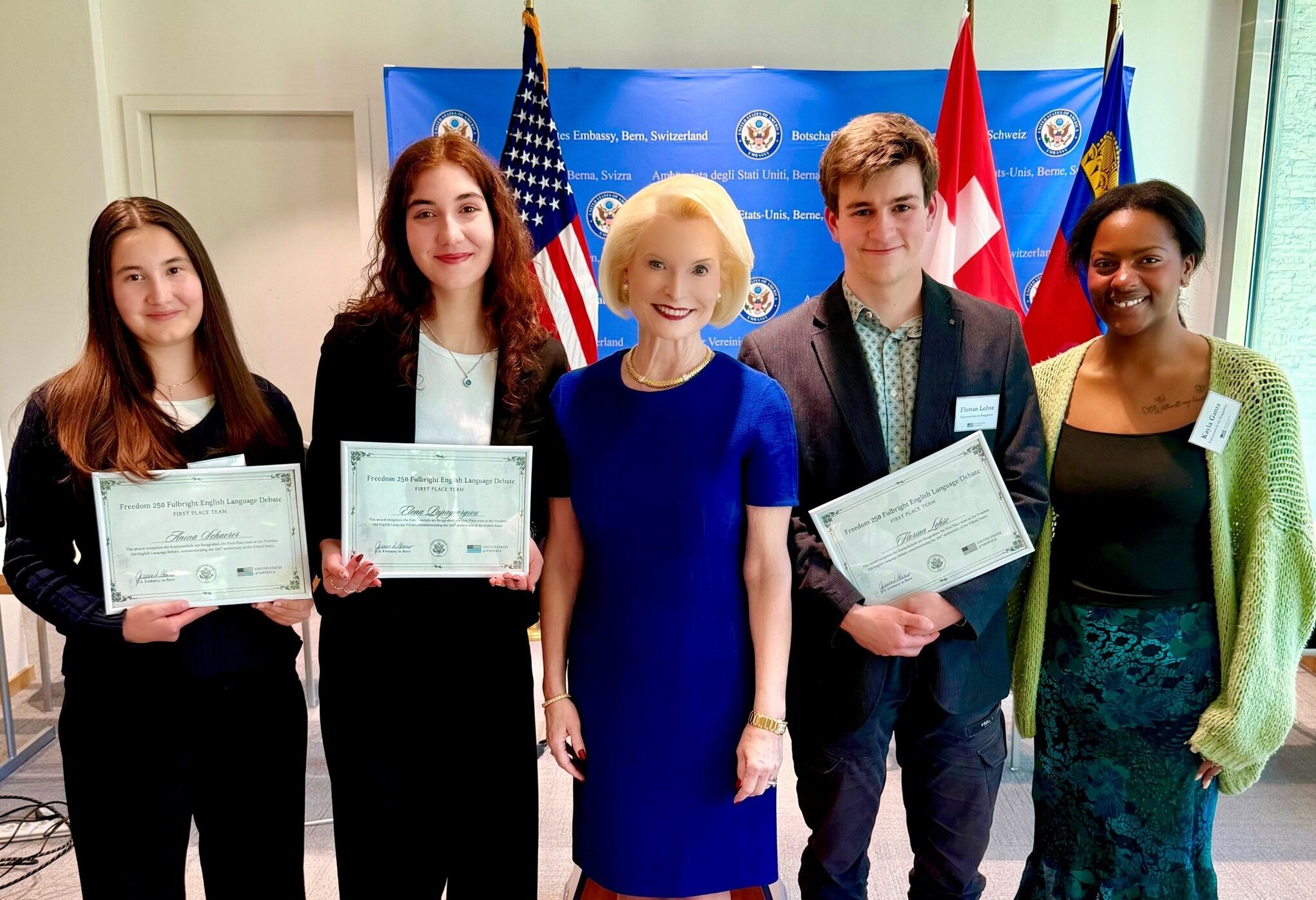 A group of four students, with three holding certificates, and standing in front of U.S. and Swiss flags, with the U.S. Ambassador to Switzerland wearing a blue dress standing in the center.