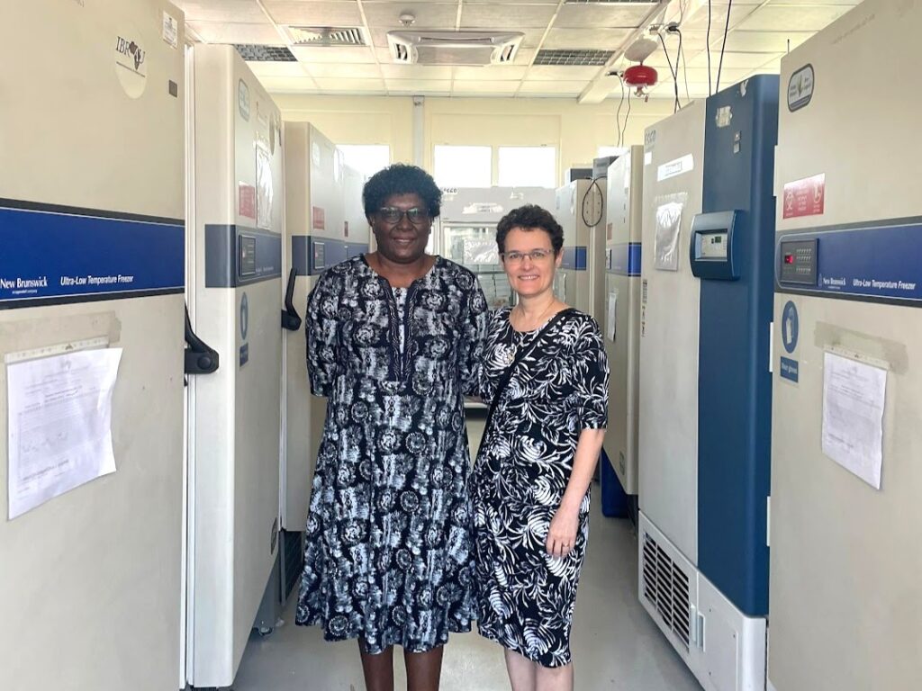Two women wearing blue patterned dresses standing in a long room with scientific refrigeration equipment on either side