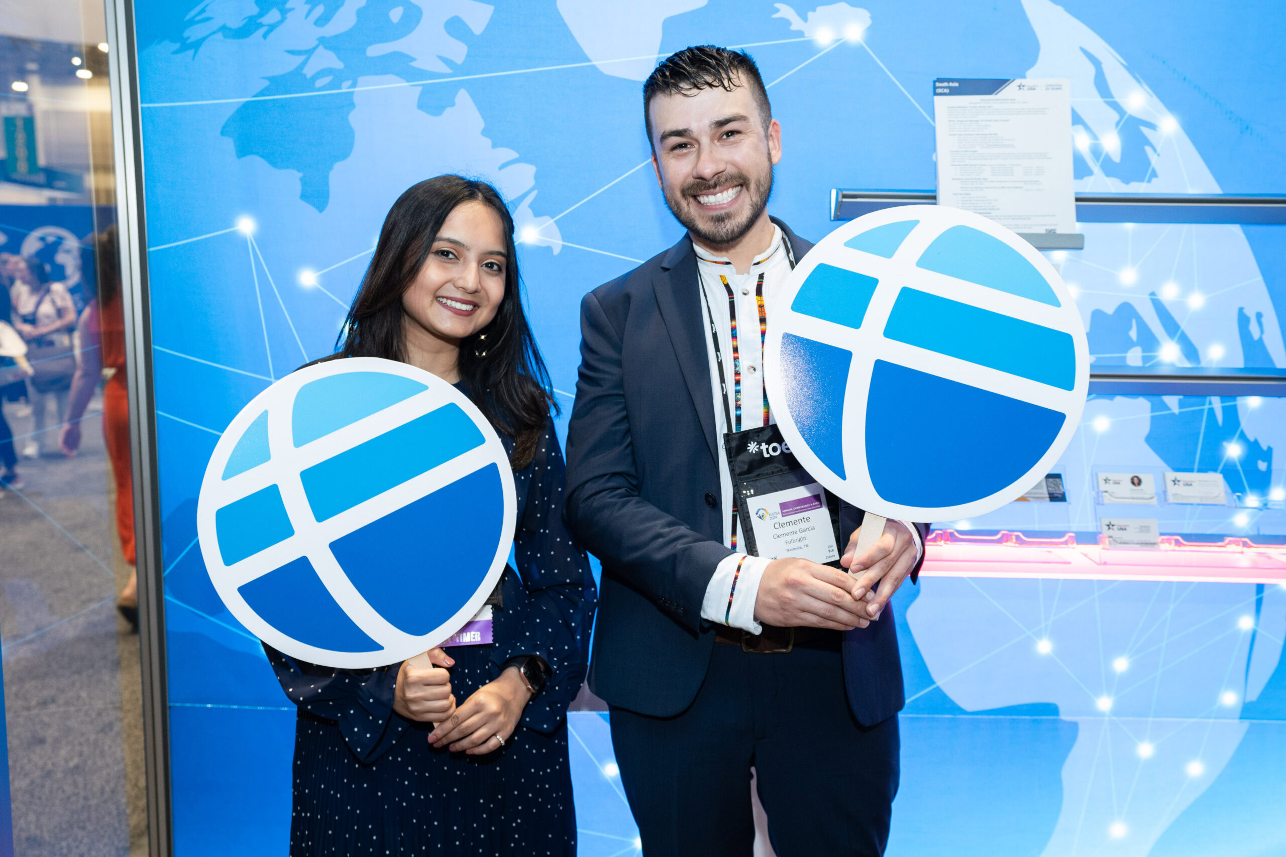 Two people wearing business attire and posing holding signs with round globe-style Fulbright logos while standing with their backs to a blue display panel with a globe graphic.