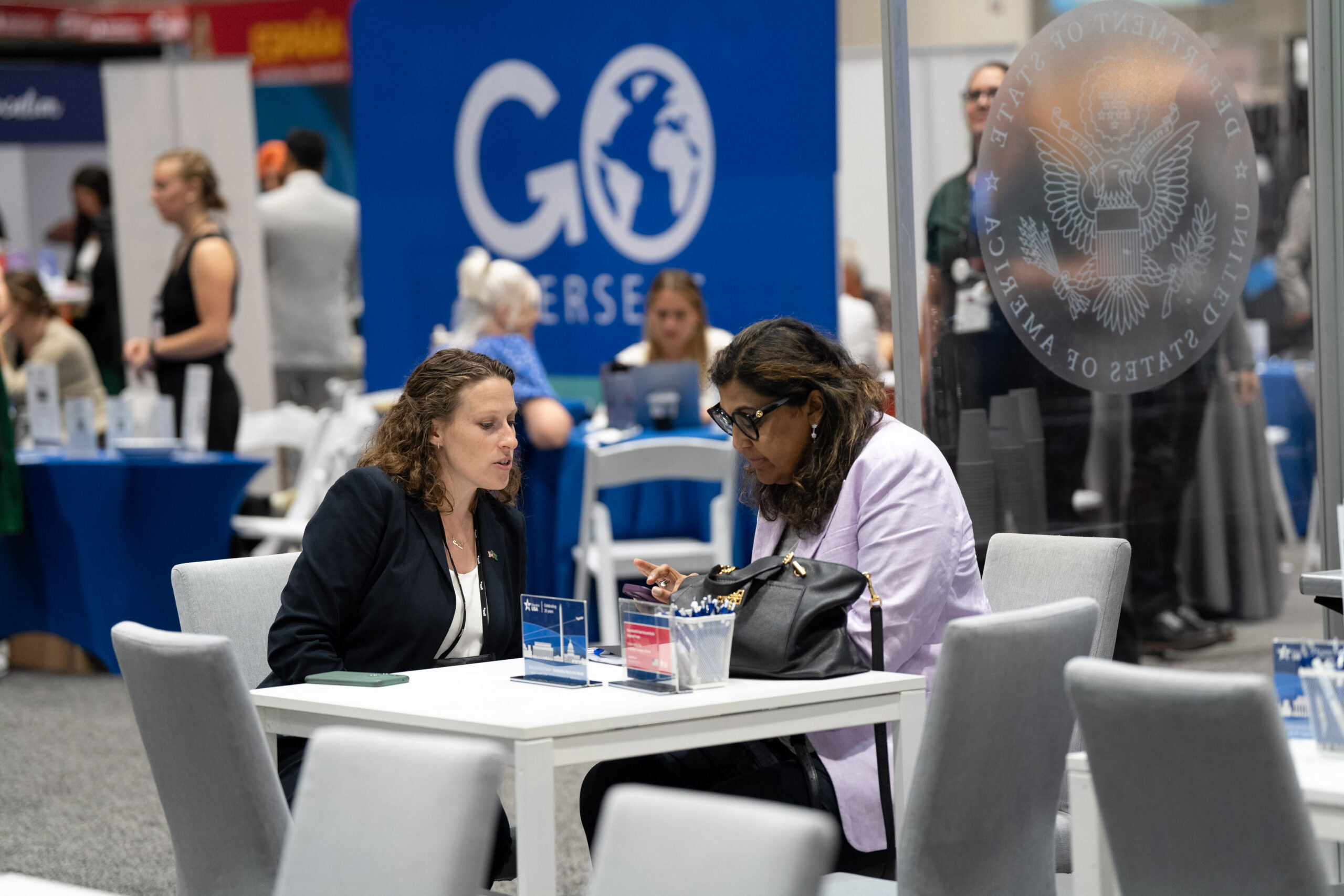 Two women wearing business attire sitting at a white table holding a meeting at an open area in an exhibit booth in a conference hall.