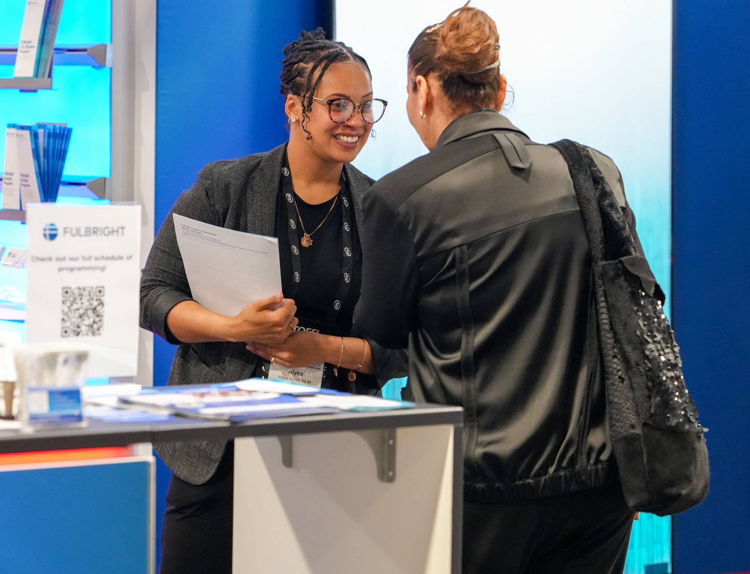 Two women wearing business attire standing next to a countertop area in an exhibit booth with blue walls and papers and brochures displayed on racks and counters.
