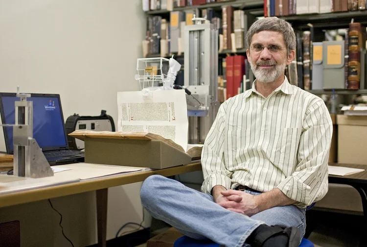 Man in striped shirt with glasses and beard sitting in front of antique bound book with a machine holding up a delicate page, in front of a shelf of antique books and manuscripts