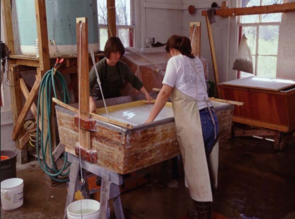 Two women wearing aprons and standing across from eachother at a large wooden mould in a workroom extracting water during the papermaking process