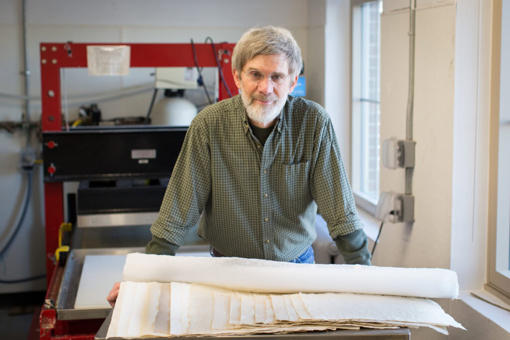 Man in green button down shirt with glasses and beard standing in front or large papermaking machine, with large sheets of hand crafted and hand cut paper in front of him