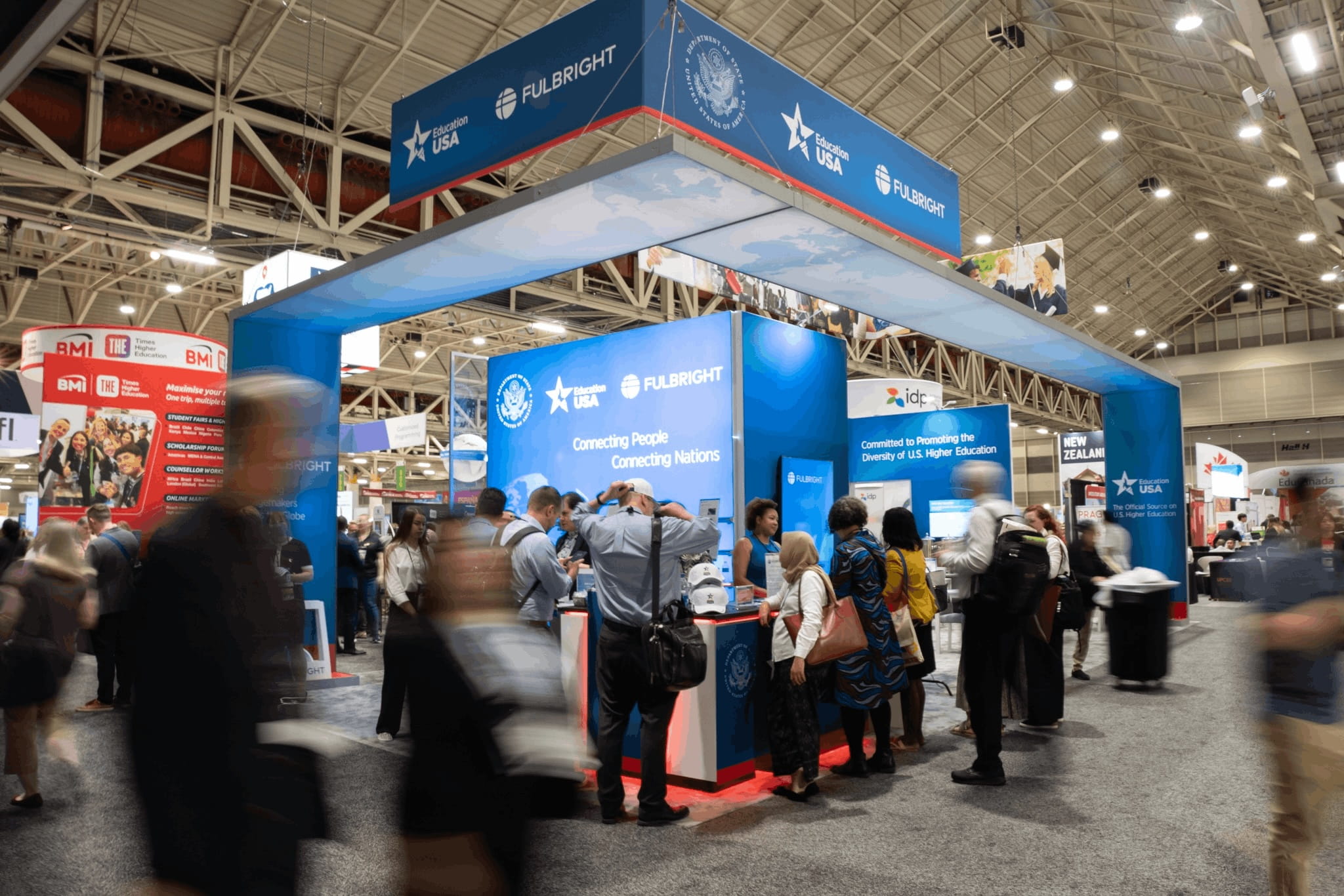 People visiting and walking past the Fulbright exhibit booth and USA Pavilion with blue and white display panels and hanging signs with Fulbright logos in a large convention center exhibit hall with high ceilings and metal beams.