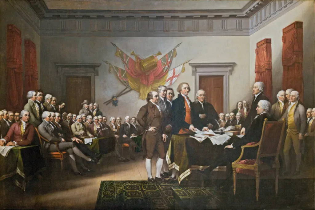 Painting of Continental Congress delegates in 1770s attire signing the Declaration of Independence by John Trumbull, showing five men in front of a large desk, with one sitting and several standing at the side, in front of rowns of men sitting and watching.