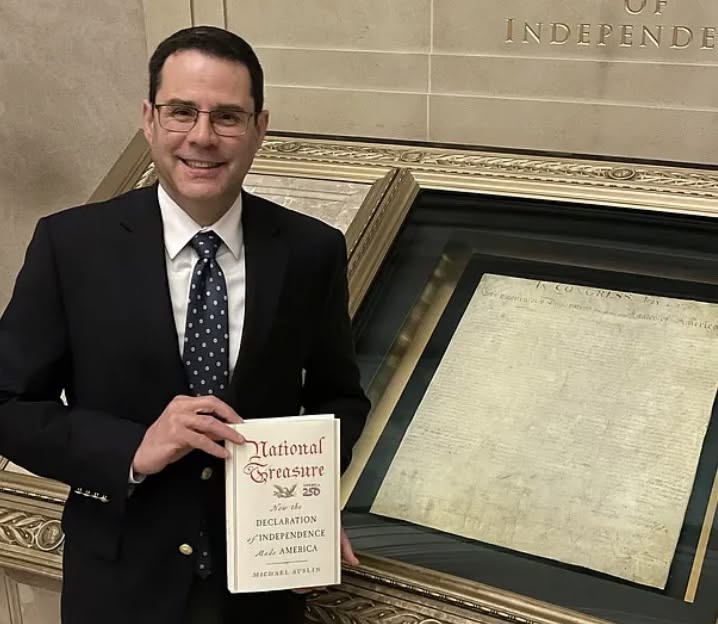 Man in blue blazer and white shirt with blue and white tie standing in front of the Declaration of Independence in its display case at the National Archives, holding a copy of a book, with the title on the front cover, saying National Treasure: Howe the Declaration of Independence Made America; Michael Auslin