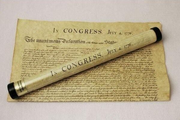 Replica of Declaration of Independence on imitation yellowed parchment paper, and a copy of it rolled up in a plastic tube, as a product displayed in a gift shop to purchase. Words showing "In Congress, July 4, 1776" at the top and calligraphied text below.