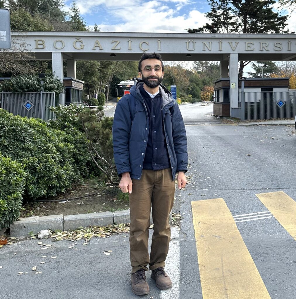 Man wearing blue jacket standing in crosswalk of street with a bridge overpass behind him that says Boğaziçi University