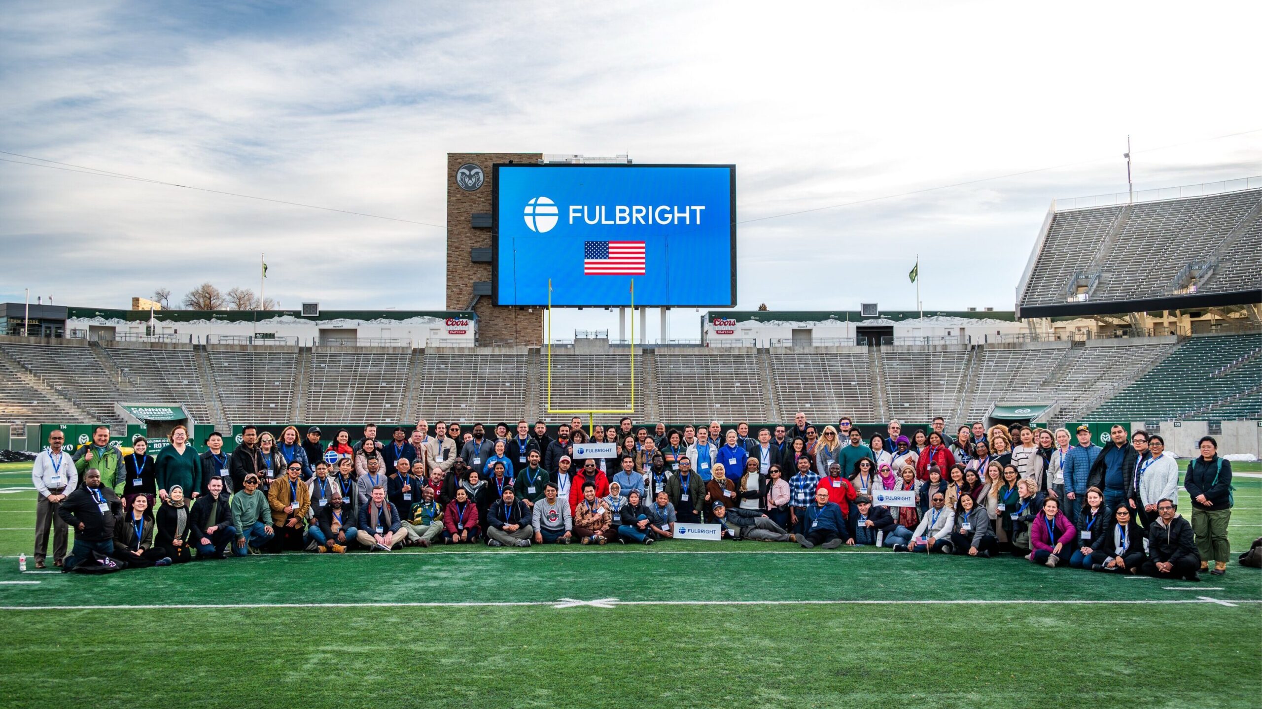 A large group of scholars sitting and standing in rows on a football field in a large stadium, with a goal post behind them and a video scoreboard showing the Fulbright logo and an American flag.