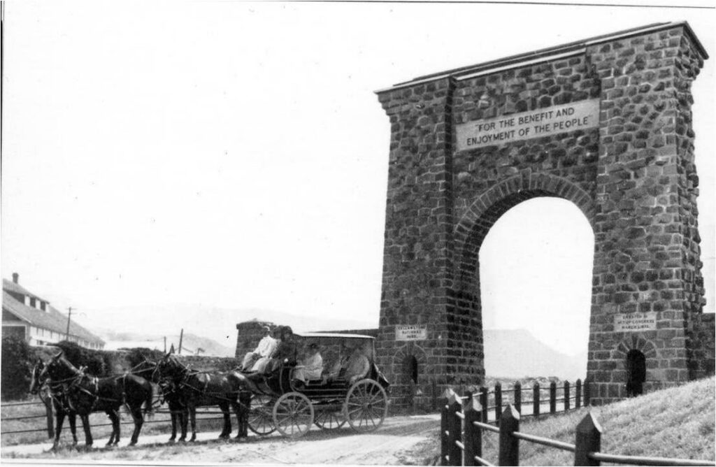 Historic scene of a horse drawn stagecoach in front of a large stone arch at Yellowstone National Park, with a stone carved plaque at the top that says For the Benefit and Enjoyment of the People. Circa 1914