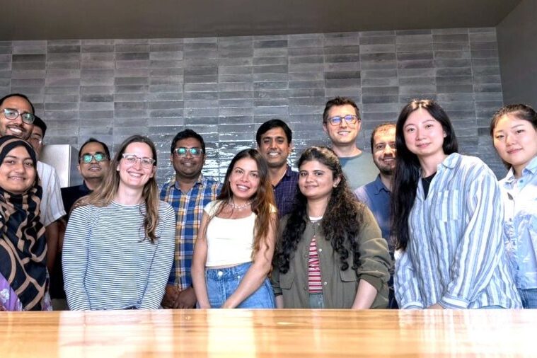 Group of 13 young adult researchers standing and smiling for a group photo in and indoor university room in front of a gray tiled wall.