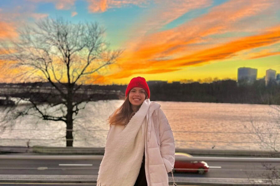 Smiling woman in winter clothing wearing a red had stands in front of the Potamac river at sunset, with a bridge, a leafless tree, and a few tall buildings behind her.