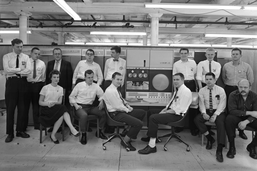 Fourteen people dressed in 1960s office attire, in two rows, sitting or standing in front of a large mainframe room-sized computer