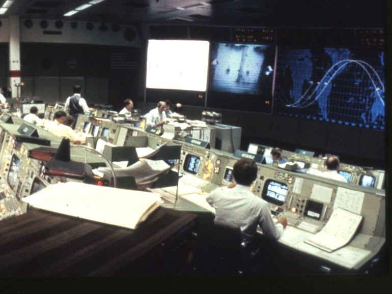 A control room at NASA showing several rows of men sitting at terminals with large display screens at the front of the room with scientific images