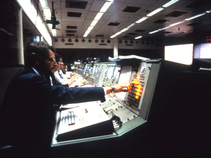 A control room at NASA with a row of men sitting in front of terminals