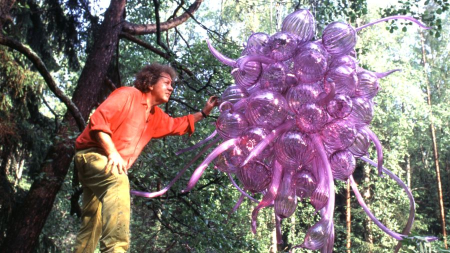 Man wearing orange shirt and yellow slacks standing in wooded setting in nature, touching and looking at large purple blown glass sculpture suspended over the landscape