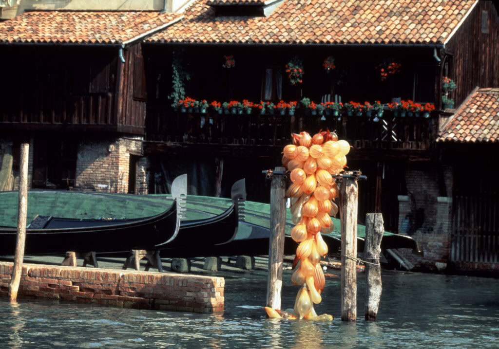 Large orange glass sculpture made out of glass bulb shapes shown suspended from pilings in a Venice canal with gondolas in the background.