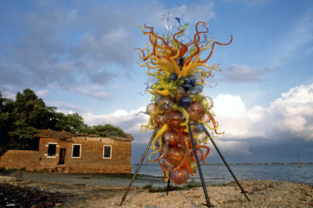 Large multicolored glass "chandelier" sculpture with varied sized bulbs and curling tentacle shapes supported by metal poles on a beach setting at the edge of the water on an island
 in Venice