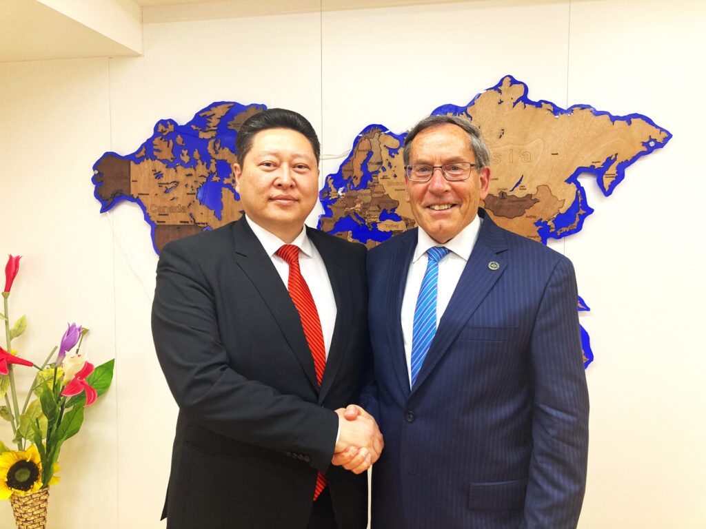 Two men shaking hands, wearing civilian suits and ties and standing in front of a ceremonial cut-out map sculptural image mounted on a wall