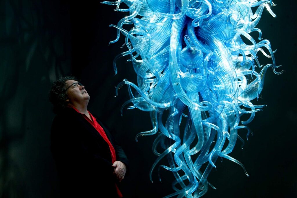 Man wearing red shirt and black coat looking up at large blue glass sculpture