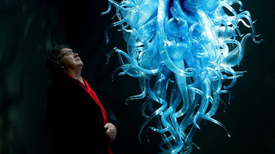 Man wearing red shirt and black coat  looking up at large blue glass sculpture 