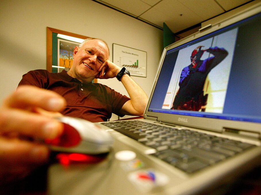 Man sitting in modern office with a Dell laptop computer open in front of him in the foreground and an image of himself on the screen.