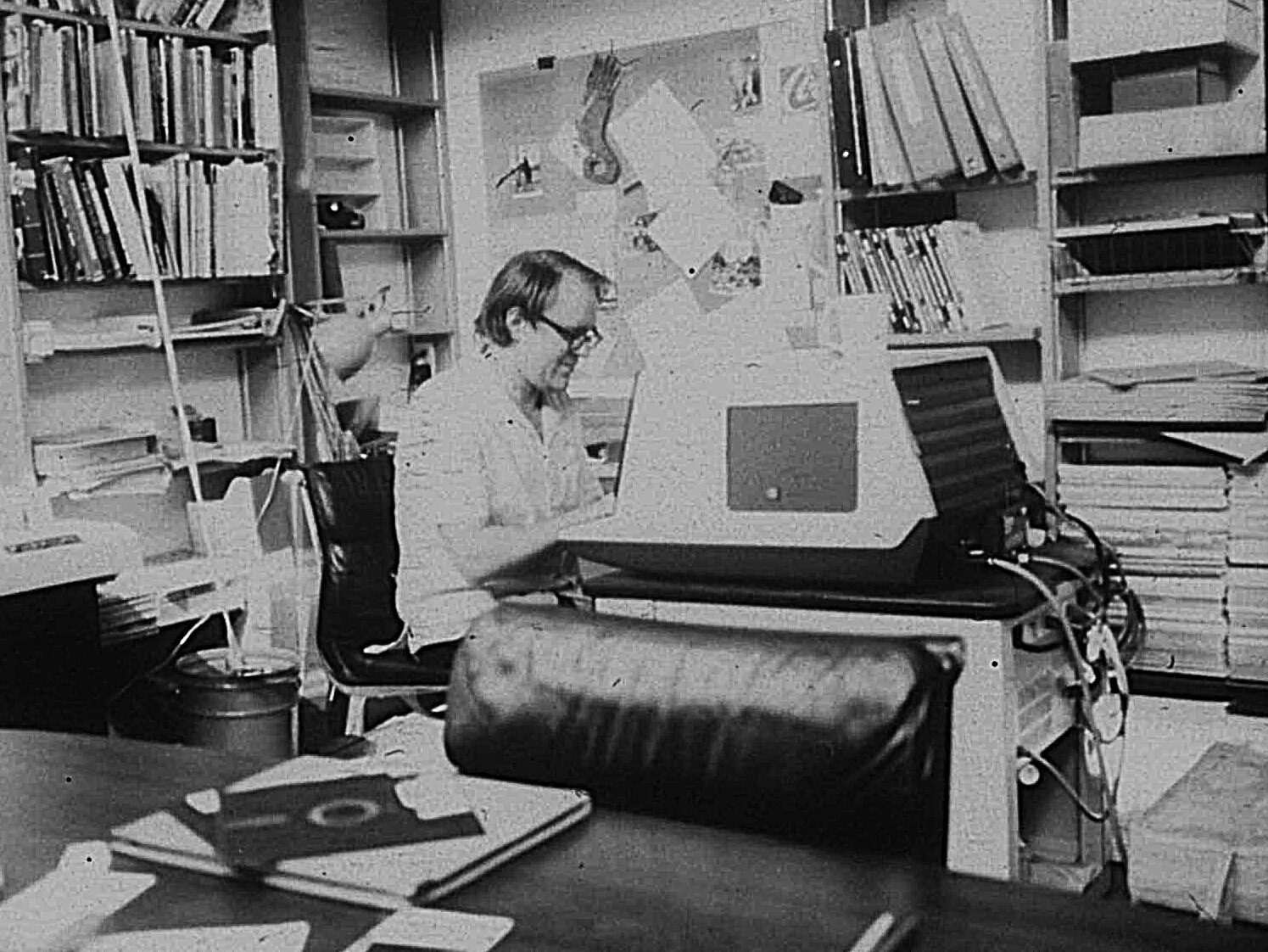 Man sitting at a large terminal on a desk surrounded by bookshelves with binders and papers, with a floppy disk on table in foreground.