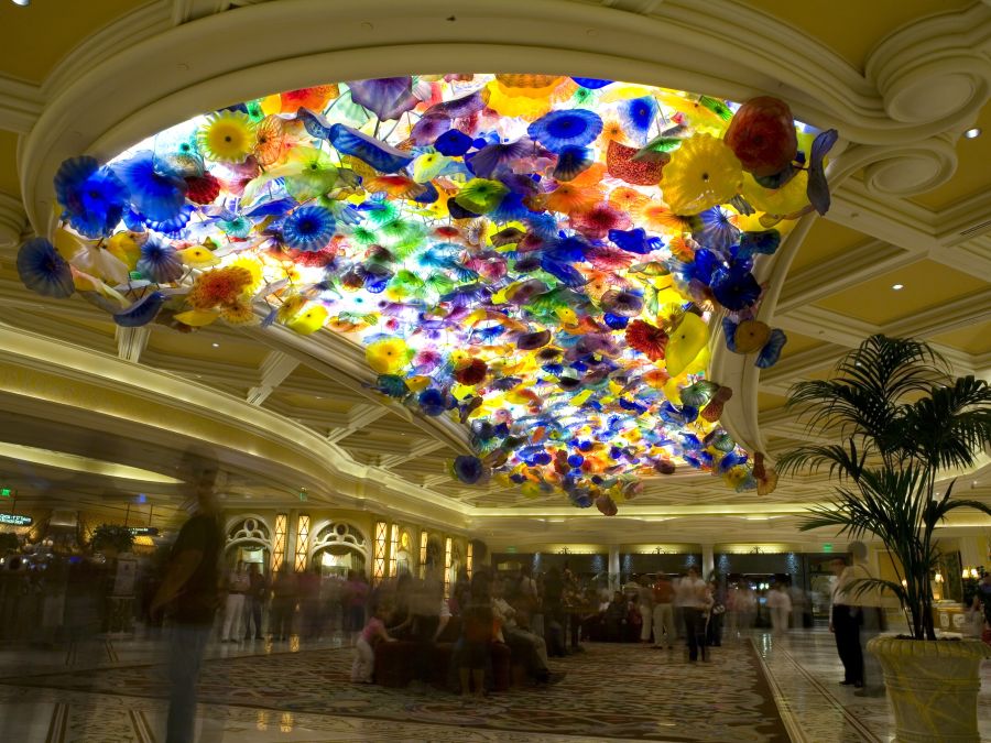 Bright multi-colored glass sculpture with flower-like shapes mounted to the ceiling of a large hotel lobby, above hotel guests and floor plants.