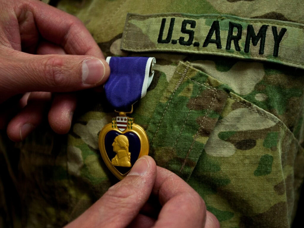 Close up view of hands pinning a heart-shaped medal with George Washington's profile in gold against a purple background, onto a U.S. Army uniform