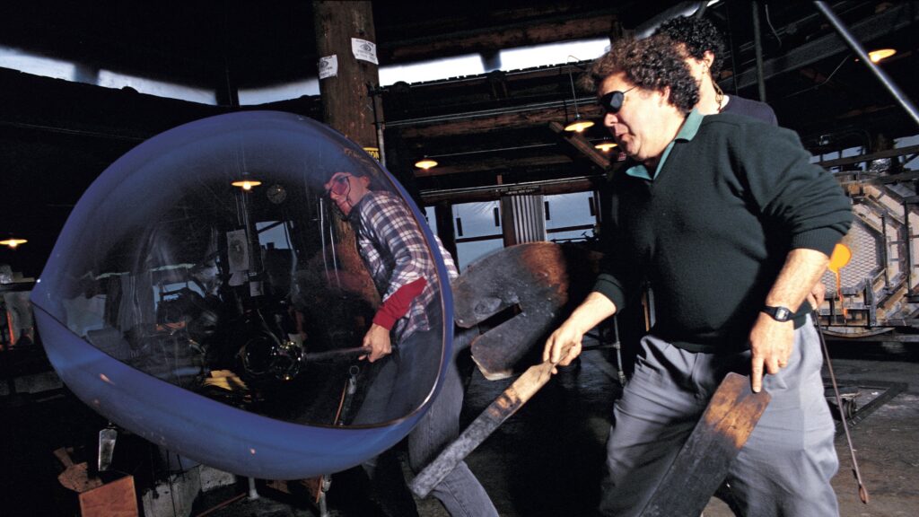 Two men working with wooden paddles and metal tools on a large blue molten glass shape in a glassmaking studio workshop