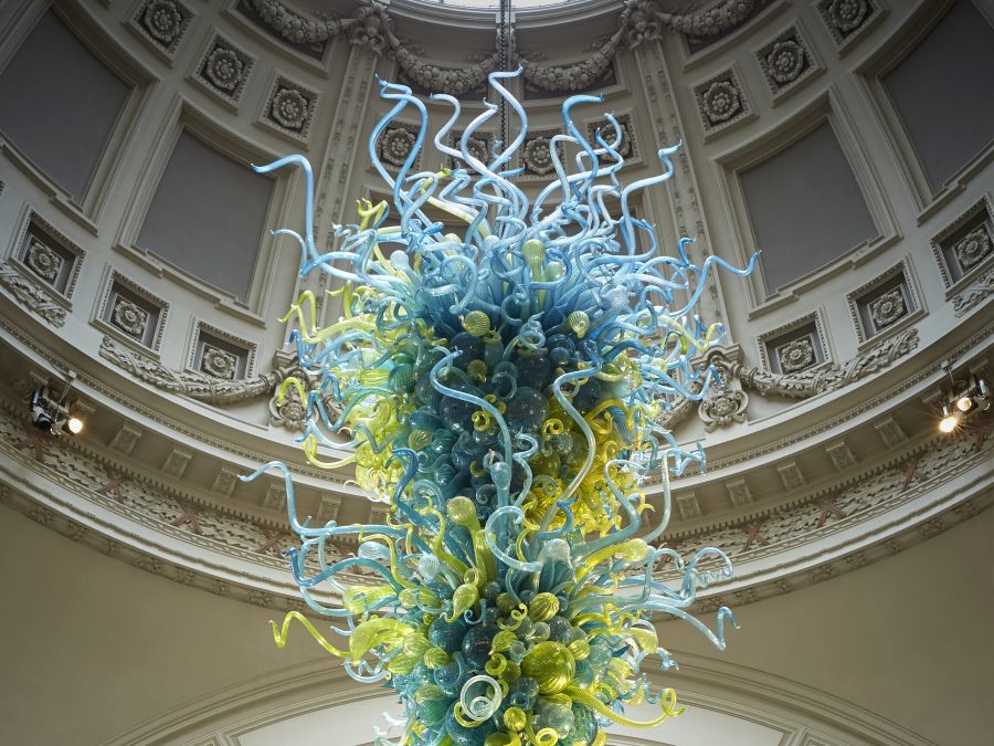 Large blue and yellow glass sculpture with glass bulb shapes and curled tentacle shapes hanging from ceiling in classical dome-shaped museum gallery room.