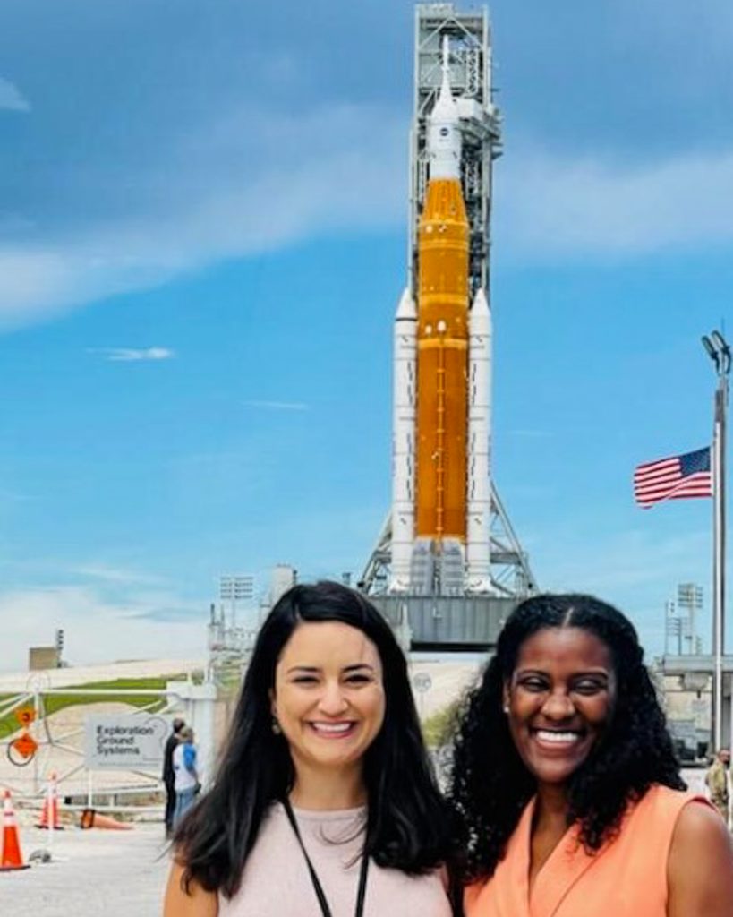 Two smiling women pose in front of NASA’s orange-and-white Artemis rocket, with an American flag and launch site structures under a blue sky.