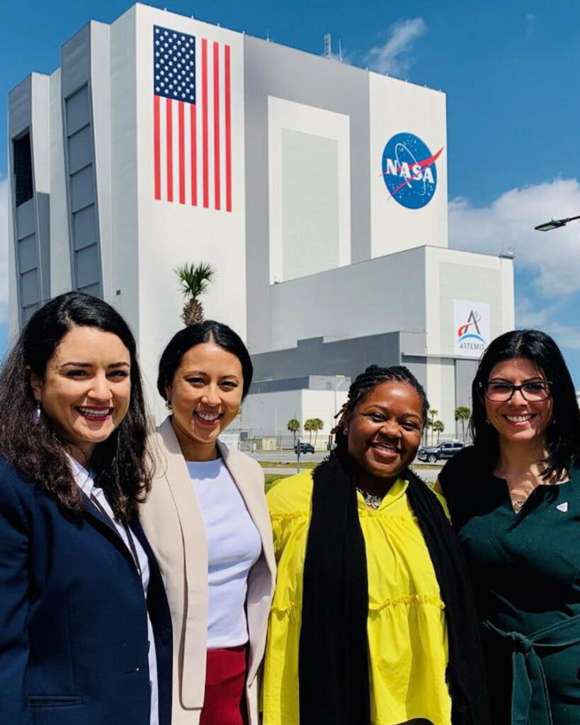 Four smiling women pose outside a NASA building beneath a large American flag and NASA logo on a bright, sunny day.