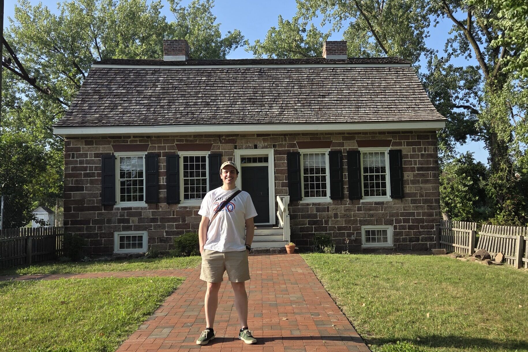 Man standing on a brick path in front of a small, historic stone house with 4 windows with shutters and two chimneys