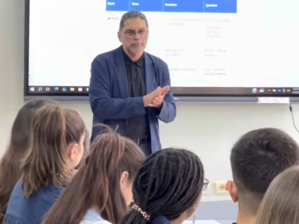 Man in suit standing in front of a screen at the front of a classroom with students facing him at a long table