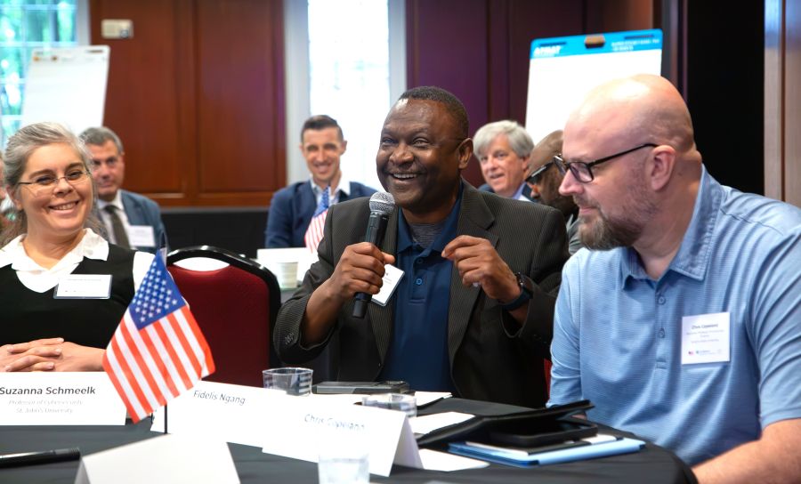 A group of people in business attire sitting at a conference table with namecards and an American flag in front of them, one holding a microphone