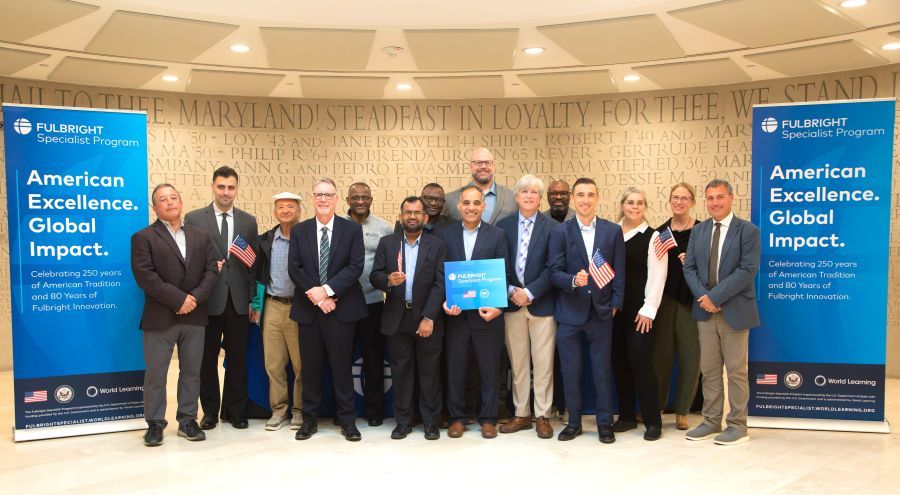 A group of 15 people in posed rows wearing business attire, standing in front of a wall inscribed with large letters with the University of Maryland motto, with blue vertical Fulbright banners at left and right of the group