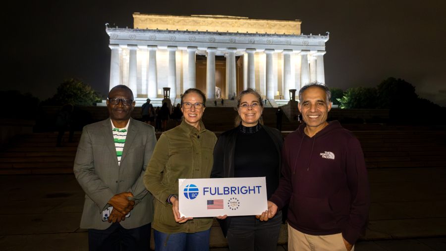 Two women and two men standing in front of a white marble monument lit up by spotlights, at night, holding a sign with Fulbright logo, American flag, and Freedom 250 logo