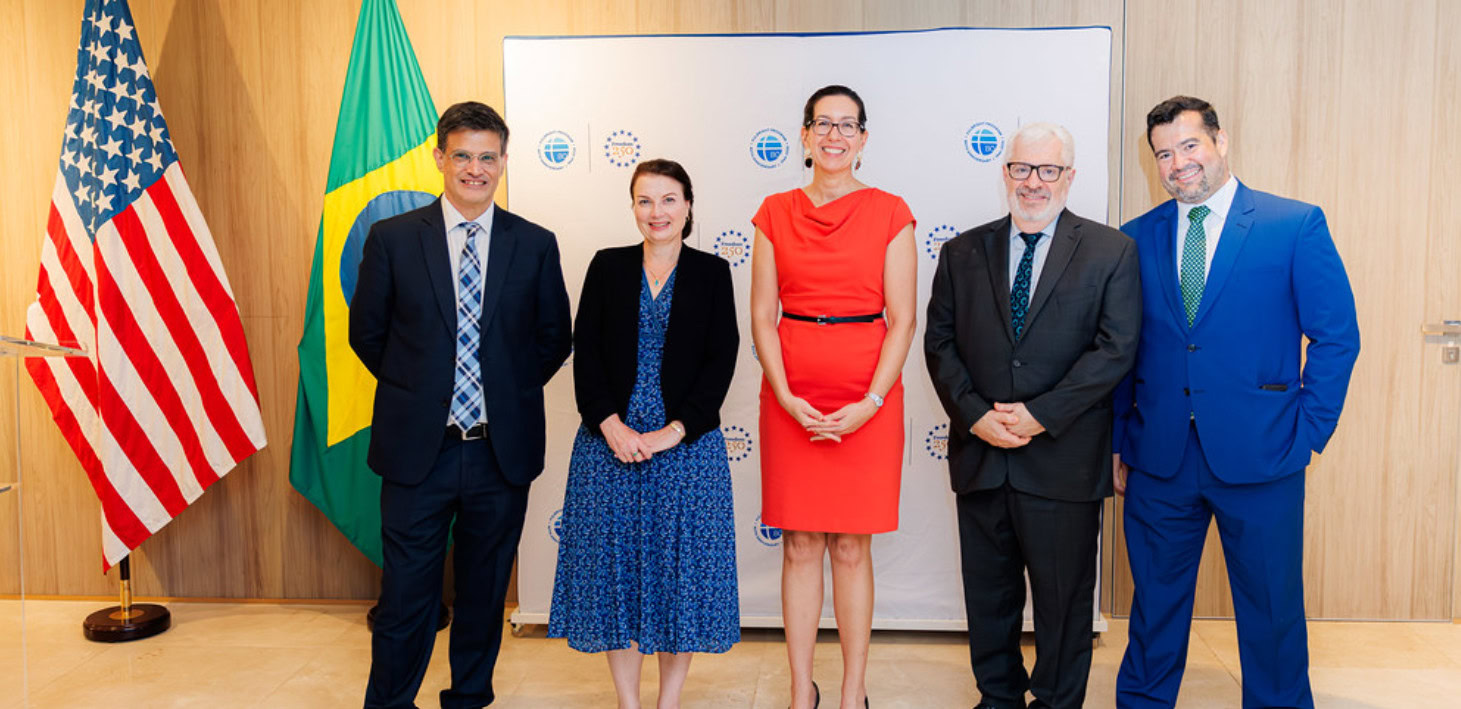 Group of five people wearing business attire standing next to Brazilian flag and American flag in front of background with Fulbright and Freedom 250 logos