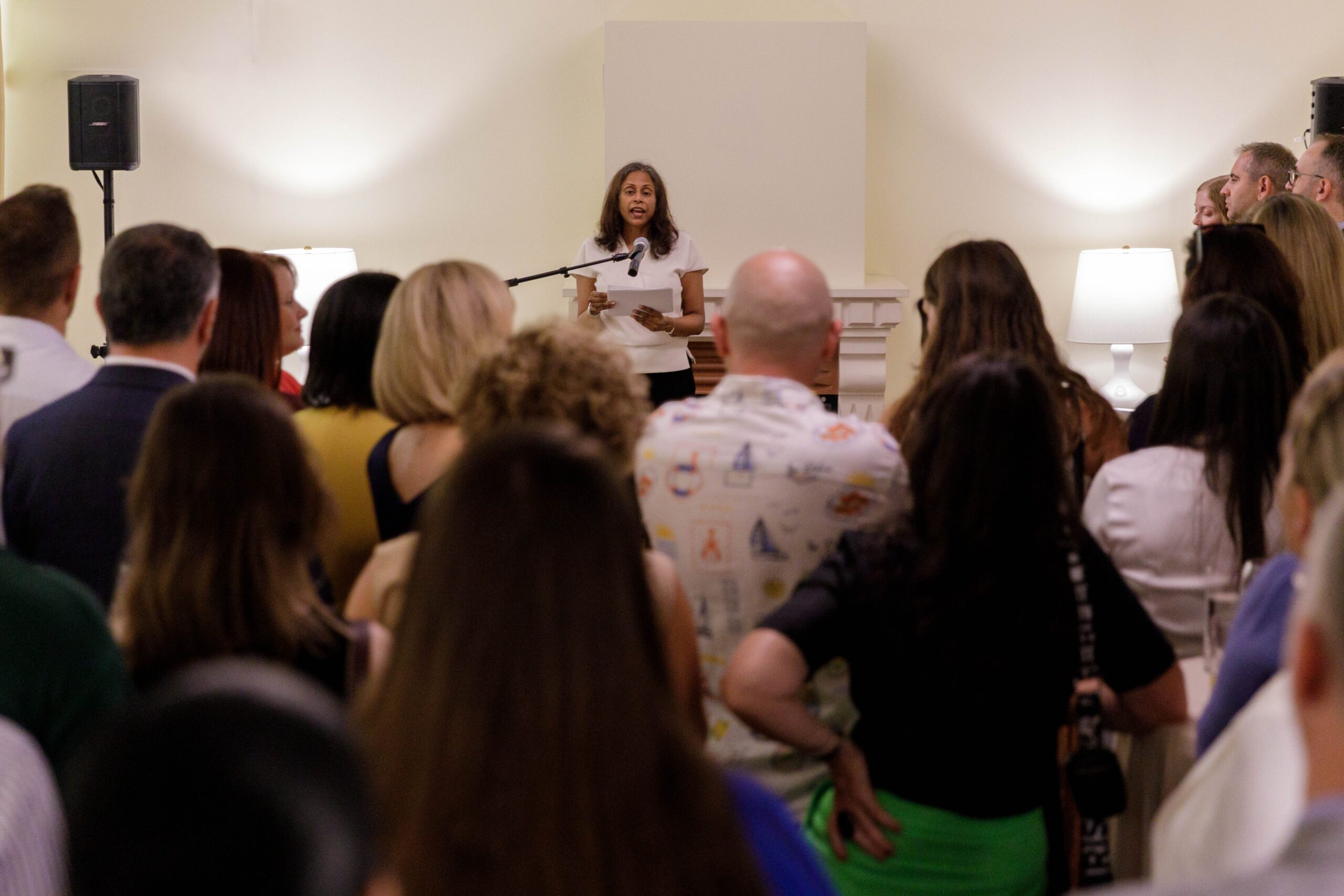 Woman standing with microphone speaking to a group of people facing her with their backs shown
