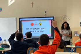 Young woman teacher standing in front of classroom with young students facing her sitting at desks raising their hands