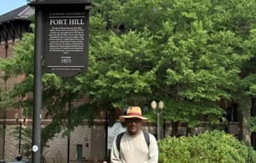 Man wearing a straw hat in front of a historic landmark building and green trees with a sign that says Fort Hill and has educational text