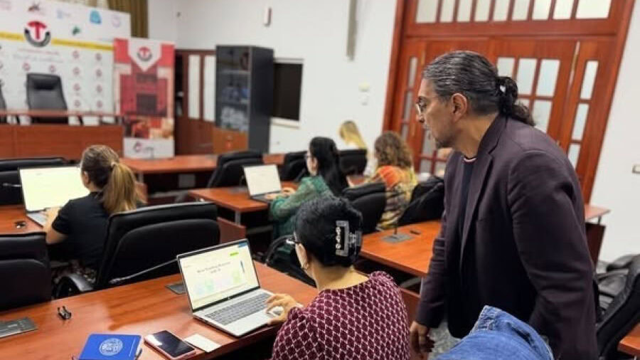 Man standing in classroom behind row of students working on laptop computers, looking at a student's work on her computer