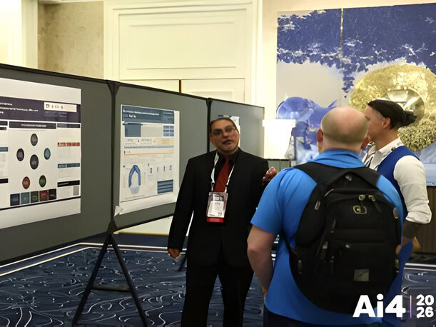 Man in suit standing in front of display board with poster talking to people gathered around him