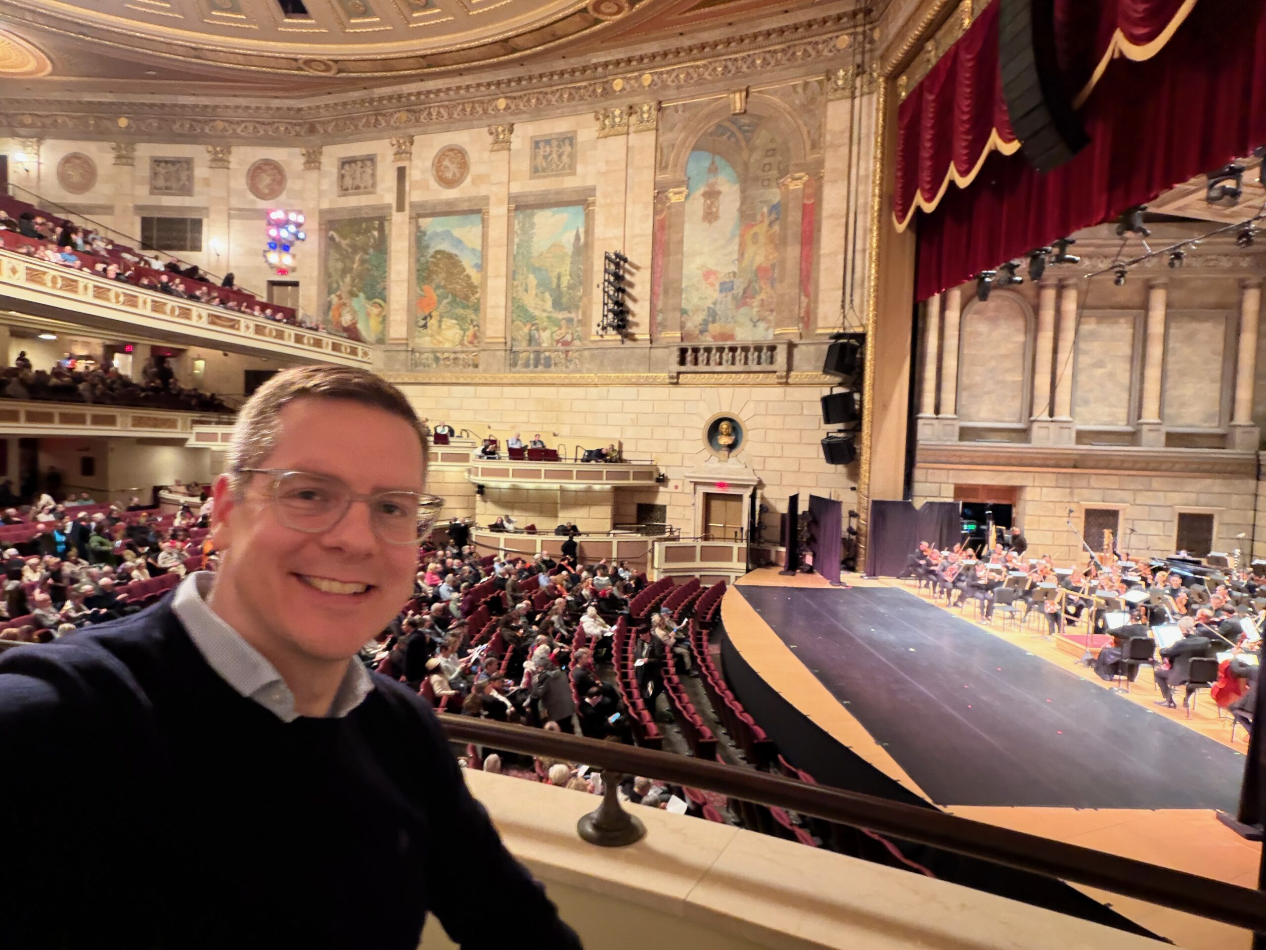 Man in an elaborate theater setting with red seats and stage curtain and murals on the walls, with orchestra musicians on the stage