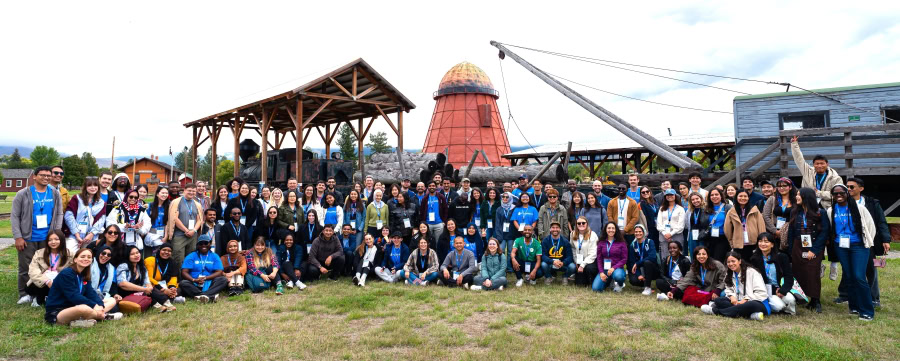Group of young professionals standing in rows in front of a restored historic train depot with large logs for lumber industry